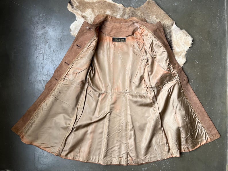 May include: Open view of a vintage brown suede jacket with a tan satin lining. The jacket has a classic collar, button closures, and a tailored fit. The lining has a label with text. The jacket is laid out on a textured surface.