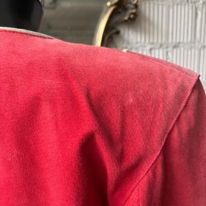 以下が含まれることがあります： 赤いスエードジャケットのクローズアップ。 ジャケットは、構造化された肩と目に見える縫い目を持つクラシックなデザインです。 生地は柔らかく、質感のある外観です。 襟は白です。