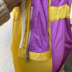 May include: Close-up of a two-tone athletic jacket in mustard yellow and purple. The jacket features a full-length zipper, a silver and gold trim, and a fitted silhouette. The fabric appears to be a lightweight, athletic material.