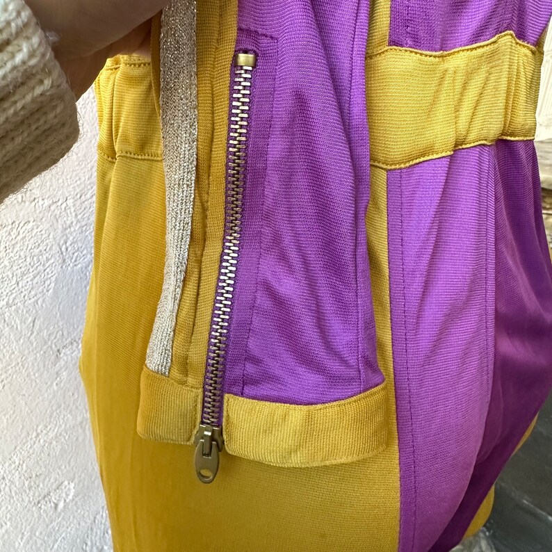 May include: Close-up of a two-tone athletic jacket in mustard yellow and purple. The jacket features a full-length zipper, a silver and gold trim, and a fitted silhouette. The fabric appears to be a lightweight, athletic material.