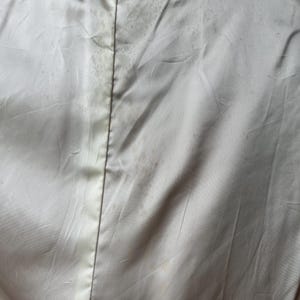 以下が含まれることがあります： 白いサテンの衣服のクローズアップ。目立つシミと中央の縫い目があります。上部にはテキスト付きのタグが付いています。生地は光沢があり、ヴィンテージ品の一部であるようです。