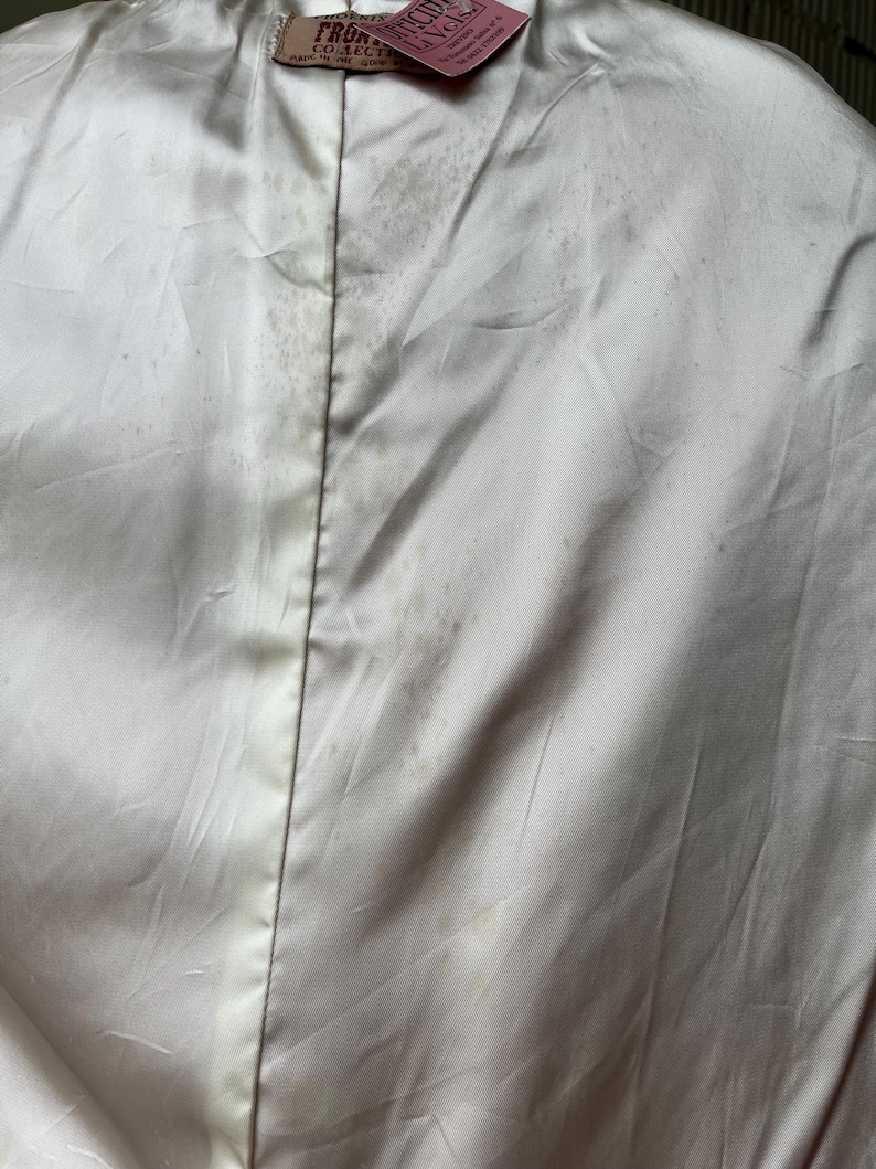 以下が含まれることがあります： 白いサテンの衣服のクローズアップ。目立つシミと中央の縫い目があります。上部にはテキスト付きのタグが付いています。生地は光沢があり、ヴィンテージ品の一部であるようです。