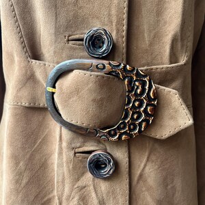 May include: Close-up of a tan suede coat with a large, decorative buckle. The buckle is a dark brown with a textured, patterned design. Two matching buttons are visible. The coat has a belt and a classic tailored design.