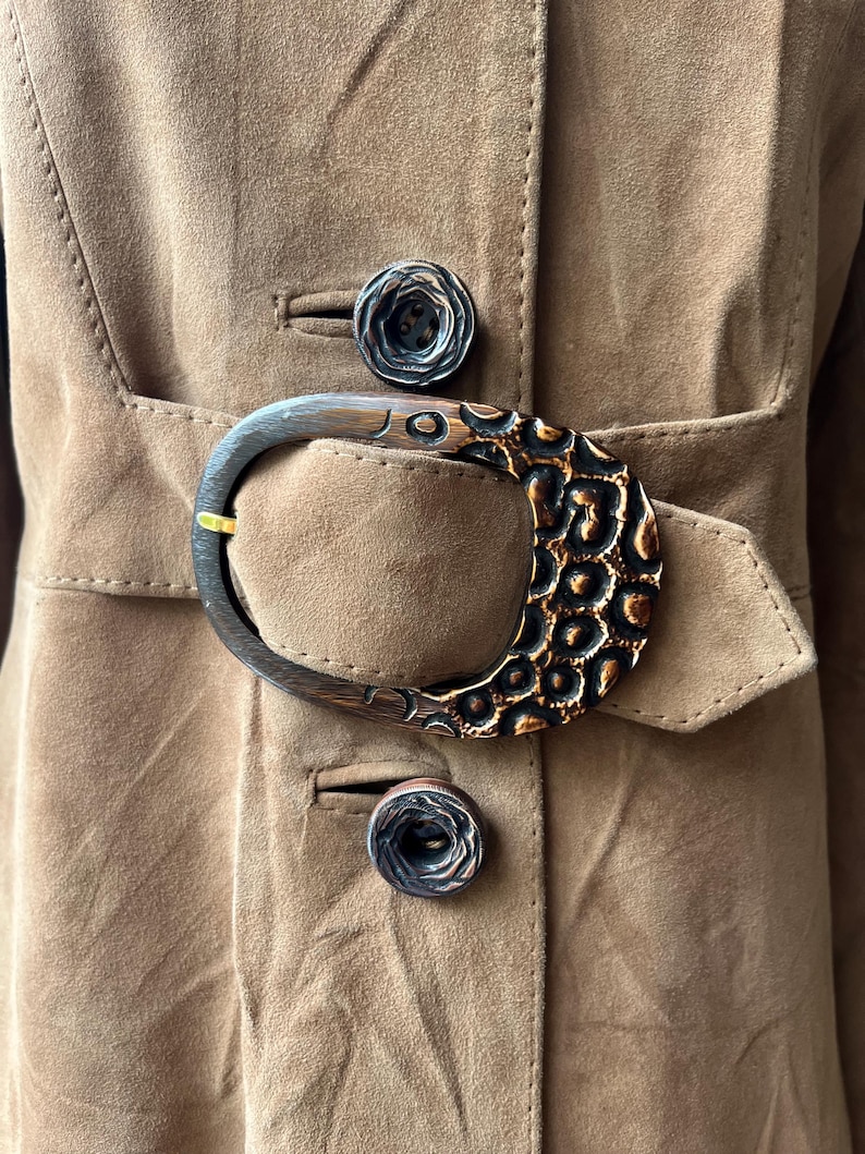May include: Close-up of a tan suede coat with a large, decorative buckle. The buckle is a dark brown with a textured, patterned design. Two matching buttons are visible. The coat has a belt and a classic tailored design.