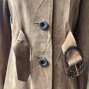 May include: A tan suede coat with a tailored fit. The coat features decorative buttons, a belt with a wooden buckle, and a classic design. The coat is a neutral color and has a vintage aesthetic.