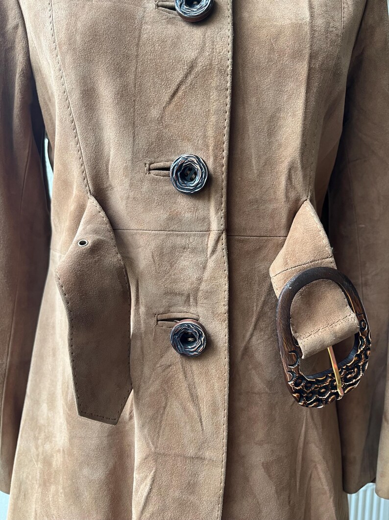 May include: A tan suede coat with a tailored fit. The coat features decorative buttons, a belt with a wooden buckle, and a classic design. The coat is a neutral color and has a vintage aesthetic.
