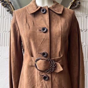 May include: A brown suede coat with a notched collar and three decorative buttons. The coat features a belt with a large, ornate buckle. The coat is displayed on a mannequin.