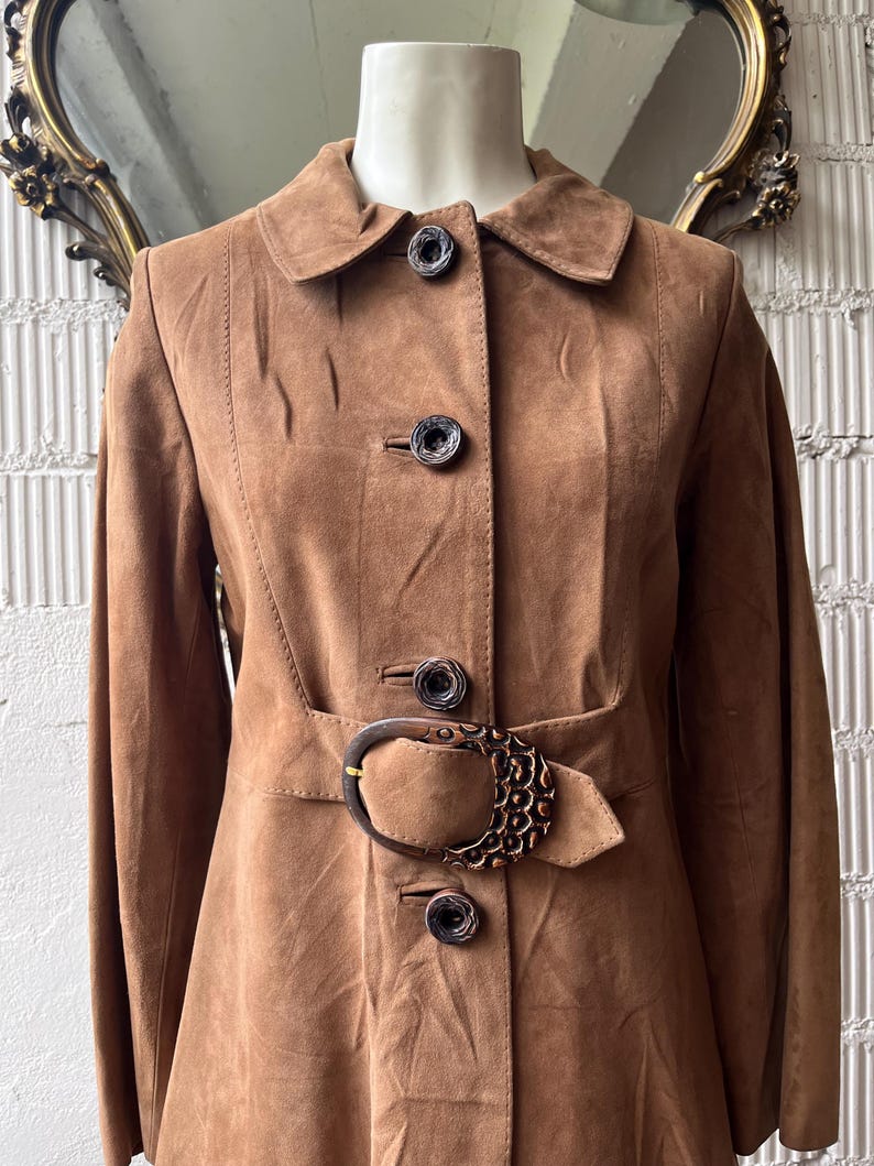 May include: A brown suede coat with a notched collar and three decorative buttons. The coat features a belt with a large, ornate buckle. The coat is displayed on a mannequin.