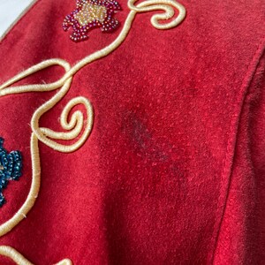 以下が含まれることがあります： 赤色のスエードの衣服のクローズアップ。赤、金、青の色合いの装飾的な花のビーズ細工が施されています。クリーム色の刺繍の渦巻きとステッチの縫い目があります。