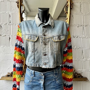 Peut inclure: Une veste en jean bleu clair avec des manches en crochet colorées. Les manches sont rayées de rouge, orange, jaune, vert, bleu et gris.
