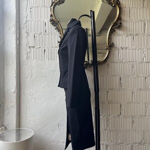 Pu&ograve; includere: Un completo nero con giacca e gonna con dettagli borchiati argentati. La giacca &egrave; sartoriale con revers e la gonna &egrave; a tubino. L'abito &egrave; appeso a un appendiabiti in metallo nero davanti a un muro di mattoni bianchi.