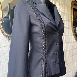 Pu&ograve; includere: Un blazer nero dalla vestibilit&agrave; sartoriale, con borchie decorative color argento lungo le cuciture e i revers. Il blazer ha un classico colletto a lancia, maniche lunghe e tre bottoni visibili. L'indumento &egrave; esposto su un manichino.
