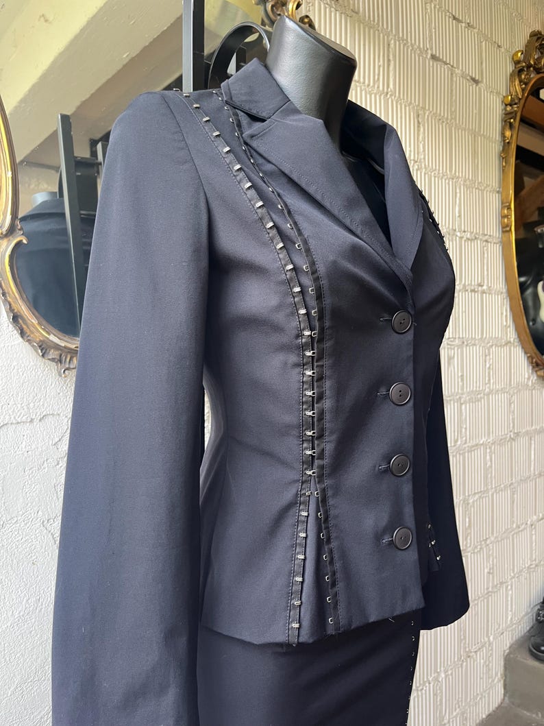 Pu&ograve; includere: Un blazer nero dalla vestibilit&agrave; sartoriale, con borchie decorative color argento lungo le cuciture e i revers. Il blazer ha un classico colletto a lancia, maniche lunghe e tre bottoni visibili. L'indumento &egrave; esposto su un manichino.