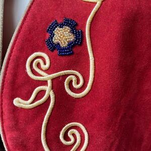 以下が含まれることがあります： クリーム色の刺繍の渦とビーズの花柄が施された赤いスエードベストのクローズアップ。花は青と金です。ベストはクリーム色のトリムが付いています。
