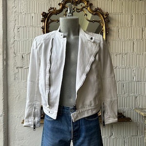 May include: A white moto jacket with zipper detailing on the sleeves and front. The jacket is on a mannequin and has a cropped length.