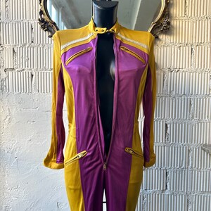 May include: Mustard yellow and purple jumpsuit with a full-length zipper. The jumpsuit features a high collar, long sleeves, and zippered pockets. Silver glitter accents the shoulders. The garment is displayed on a black mannequin.