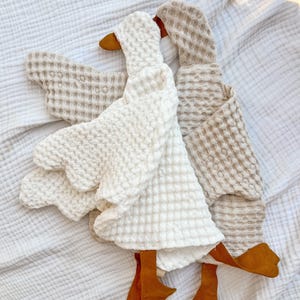 May include: Two goose-shaped plush toys made from waffle-textured fabric. One goose is white and the other is beige. Both feature brown fabric beaks and webbed feet. The toys have a soft, quilted appearance with distinct wing and neck shapes.