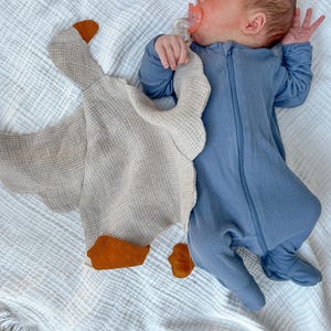 May include: A newborn baby wearing a blue ribbed onesie lies on a white textured blanket, next to a beige goose-shaped security blanket with orange accents. The baby is using a pacifier.
