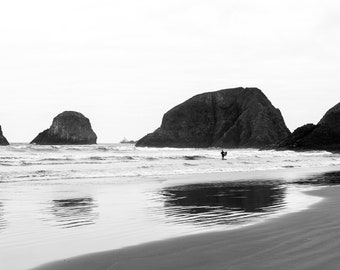 Op de afbeelding: Een zwart-witfoto van een strand met rotsachtige uitsteeksels en een eenzame surfer in het water.