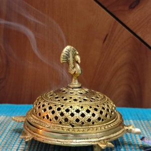 May include: A gold metal incense burner with a peacock design on the top and a decorative, openwork dome. The burner is shaped like a turtle and has three legs. Smoke is rising from the burner.