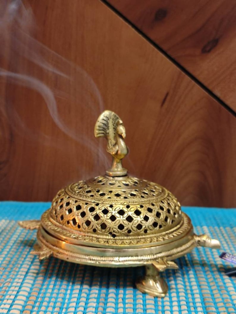 May include: A gold metal incense burner with a peacock design on the top and a decorative, openwork dome. The burner is shaped like a turtle and has three legs. Smoke is rising from the burner.