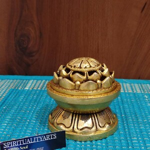 May include: A gold-colored metal incense burner shaped like a lotus flower. The burner has a round base and a top with a circular opening. The burner is sitting on a blue and white patterned surface. The text "SPIRITUALITYARTS Art With Soul HOME DECOR SPIRITUAL IDOLS" is visible on a white card in the bottom right corner of the image.