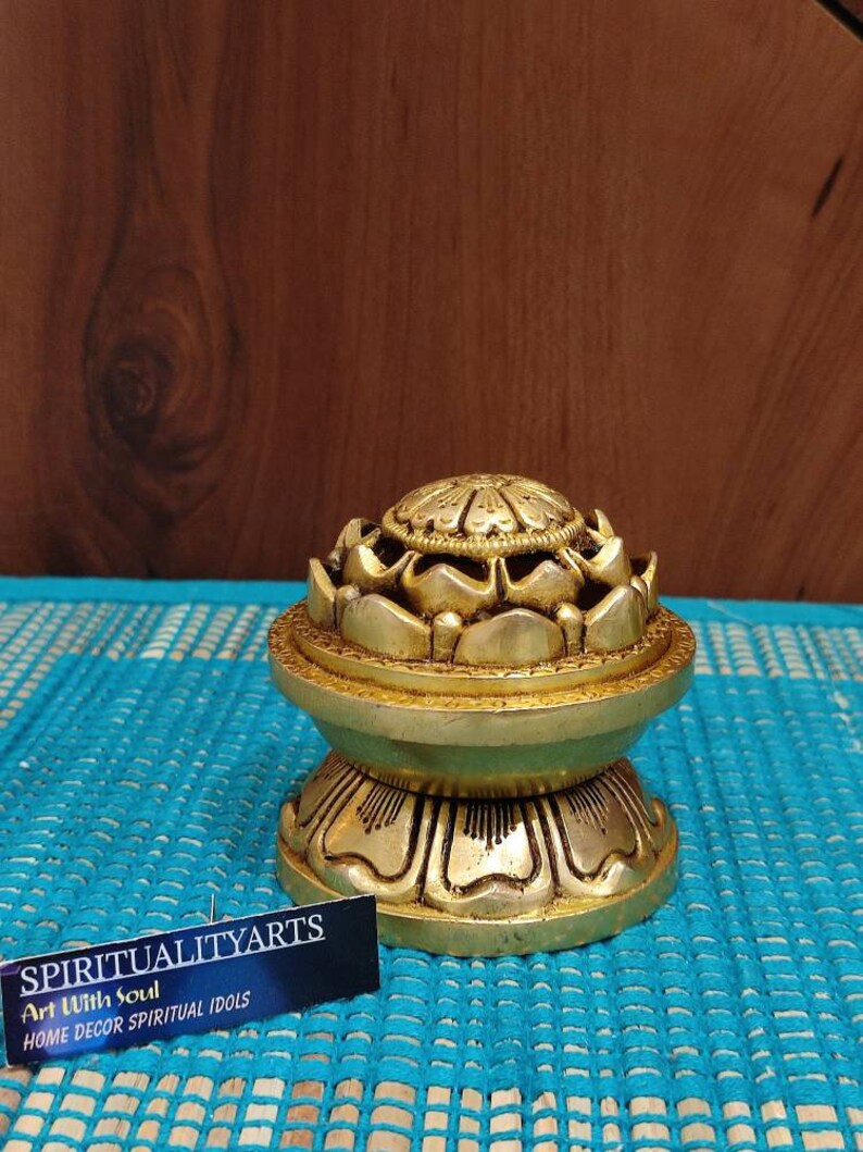 May include: A gold-colored metal incense burner shaped like a lotus flower. The burner has a round base and a top with a circular opening. The burner is sitting on a blue and white patterned surface. The text "SPIRITUALITYARTS Art With Soul HOME DECOR SPIRITUAL IDOLS" is visible on a white card in the bottom right corner of the image.