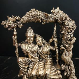 K&ouml;nnte beinhalten: Eine goldfarbene Statue eines Paares, das auf einer Schaukel sitzt. Die Schaukel ist an einem kunstvoll geschnitzten Ast eines Baumes befestigt. Das Paar ist mit aufwendigen Schmuck und traditioneller Kleidung geschm&uuml;ckt. Die Statue stellt die hinduistischen Gottheiten Radha und Krishna dar.