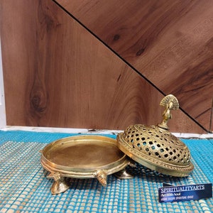 May include: A gold metal incense burner with a peacock design on the lid. The burner has a round base with four legs and a decorative design. The lid has a lattice pattern and is open. The burner is sitting on a blue and white mat. The text "Spiritualityarts Art With Soul Home Decor Spiritual Idols" is on a black card.