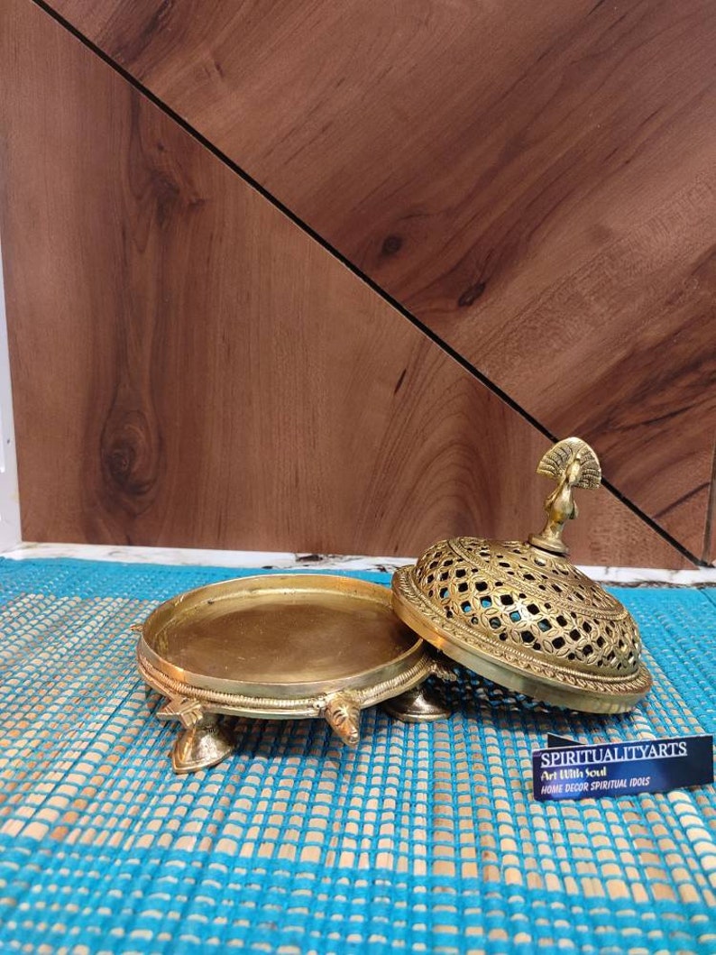 May include: A gold metal incense burner with a peacock design on the lid. The burner has a round base with four legs and a decorative design. The lid has a lattice pattern and is open. The burner is sitting on a blue and white mat. The text "Spiritualityarts Art With Soul Home Decor Spiritual Idols" is on a black card.