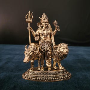 May include: A gold-colored metal statue of the Hindu deity Shiva, standing on a base with a bull and a lion on either side. Shiva is holding a trident in his right hand and a small object in his left hand.