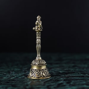 May include: A small, ornate, gold-colored bell with a detailed design. The bell has a figure of a person sitting on top of it.