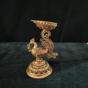 May include: A gold metal peacock-shaped diya with intricate details. The diya has a round base and a peacock design with a raised tail. The diya is perfect for adding a touch of elegance to your home decor.