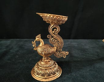May include: A gold metal peacock-shaped diya with intricate details. The diya has a round base and a peacock design with a raised tail. The diya is perfect for adding a touch of elegance to your home decor.