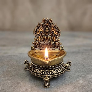 Pode incluir: Uma lâmpada a óleo em tom dourado metálico com uma placa traseira em relevo intrincada e uma figura central sentada. A taça está cheia de óleo com um pavio aceso, apoiada por três pernas ornamentadas com pequenos detalhes animais.