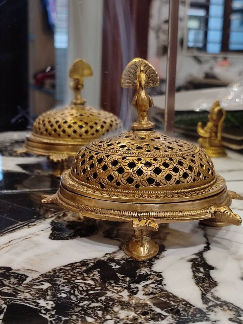 May include: A gold metal incense burner with a peacock design on the top. The burner has a round, openwork base and a domed top. The burner is sitting on a marble surface.