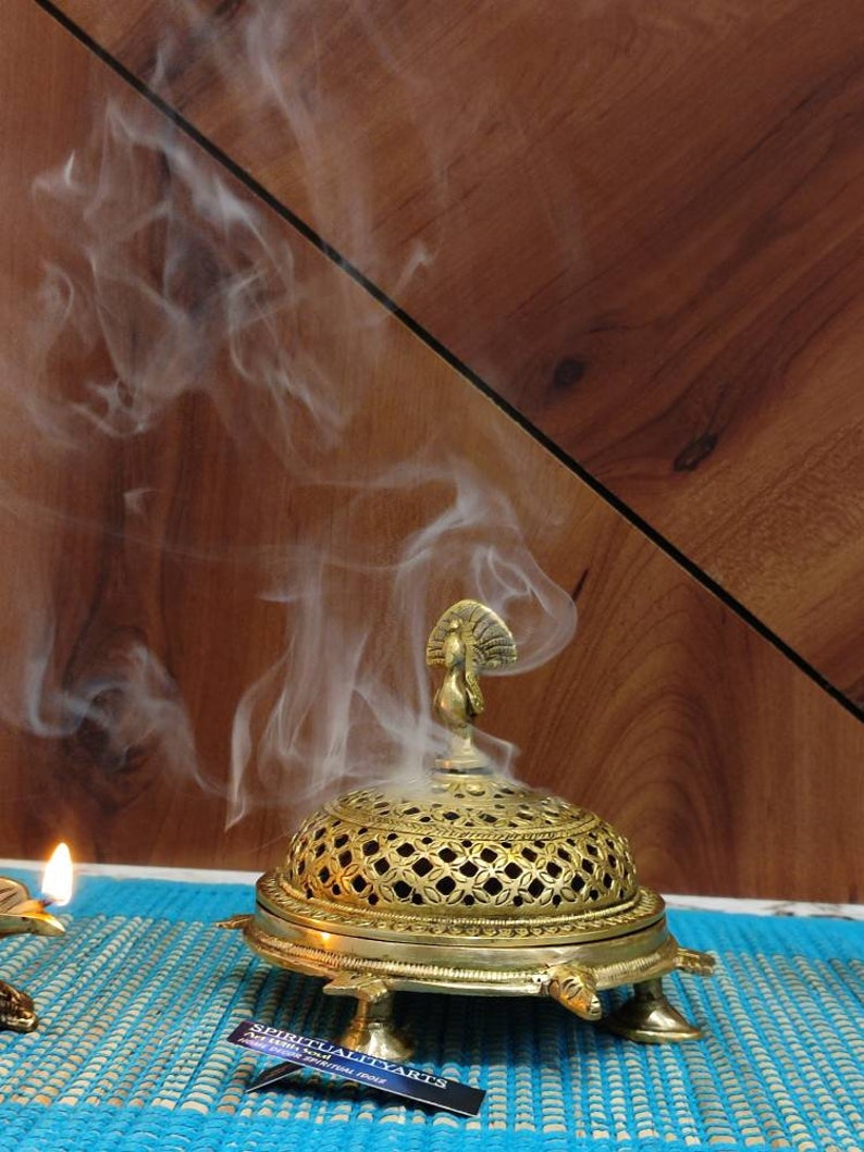 May include: A gold metal incense burner shaped like a turtle with a peacock finial on top. The burner has a decorative, openwork design and is sitting on a blue and white patterned surface. Smoke is rising from the burner.