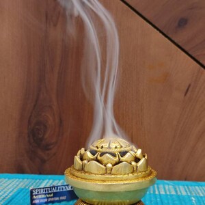 May include: A gold incense burner shaped like a lotus flower with a black center. Smoke rises from the burner, which is sitting on a blue surface. The text "SPIRITUALITY" is visible on a black and white sticker.