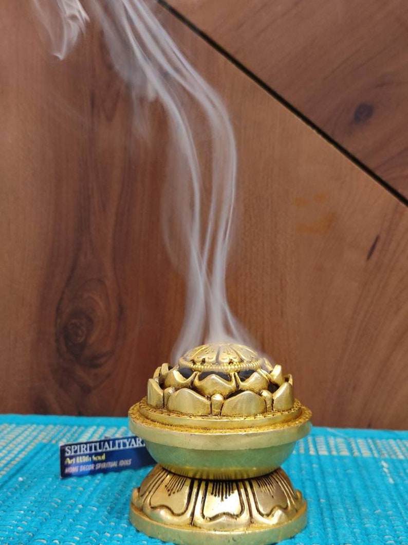 May include: A gold incense burner shaped like a lotus flower with a black center. Smoke rises from the burner, which is sitting on a blue surface. The text "SPIRITUALITY" is visible on a black and white sticker.
