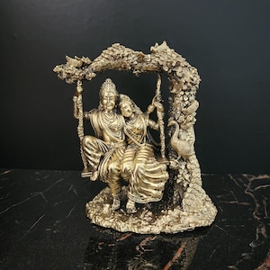 K&ouml;nnte beinhalten: Eine goldfarbene Statue eines Paares, das auf einer Schaukel unter einem Baum sitzt. Das Paar ist in traditioneller indischer Kleidung gekleidet und der Baum ist mit Blumen und Bl&auml;ttern geschm&uuml;ckt. Ein Pfau sitzt auf einem Ast des Baumes.