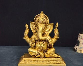 May include: A gold-colored brass statue of Ganesha, the Hindu god of wisdom and prosperity, sitting in a meditative pose with his trunk raised. The statue is detailed with intricate carvings and is placed on a rectangular base.