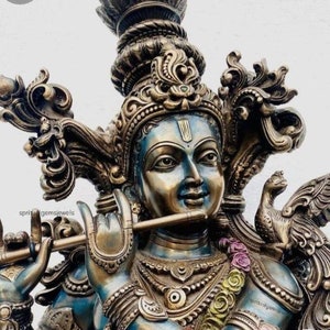 May include: A detailed, ornate statue of Krishna playing a flute. The statue is made of brass and has a blue and gold finish. Krishna is wearing a crown and jewellery, and a peacock is perched on his shoulder.
