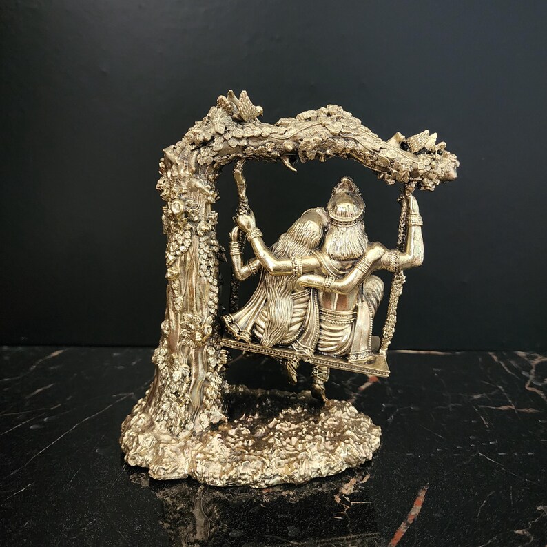 K&ouml;nnte beinhalten: Eine goldfarbene Metallskulptur eines Paares, das auf einer Schaukel sitzt. Die Schaukel ist an einem Ast mit aufwendigen Details befestigt. Die Skulptur steht auf einem Sockel mit einer strukturierten Oberfl&auml;che.