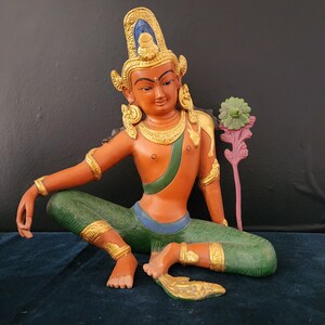 May include: A colorful statue of a deity sitting in a meditative pose. The statue is made of wood and painted in shades of orange, green, blue, and gold. The deity has a crown and holds a flower in one hand.