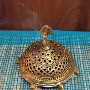 May include: A gold metal incense burner shaped like a turtle with a peacock design on top. Smoke is rising from the burner. The burner is on a blue and white patterned surface.  "SPIRITUALITYARTS Art With Soul HOME DECOR SPIRITUAL IDOLS" is printed on a black card in the bottom right corner of the image.