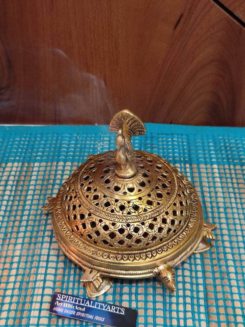 May include: A gold metal incense burner shaped like a turtle with a peacock design on top. Smoke is rising from the burner. The burner is on a blue and white patterned surface.  "SPIRITUALITYARTS Art With Soul HOME DECOR SPIRITUAL IDOLS" is printed on a black card in the bottom right corner of the image.