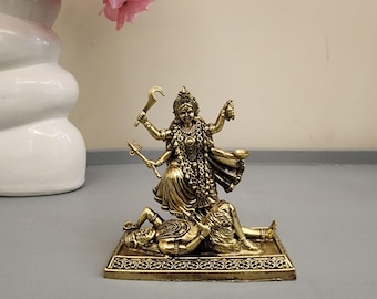 May include: A metallic gold-toned figurine depicting the Hindu goddess Kali standing over the reclining figure of Shiva. The statue features intricate detailing on the deity's multiple arms, traditional ornaments, and the rectangular base.