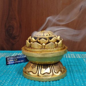 May include: A gold metal incense burner shaped like a lotus flower with a round base. Smoke is rising from the top of the burner.