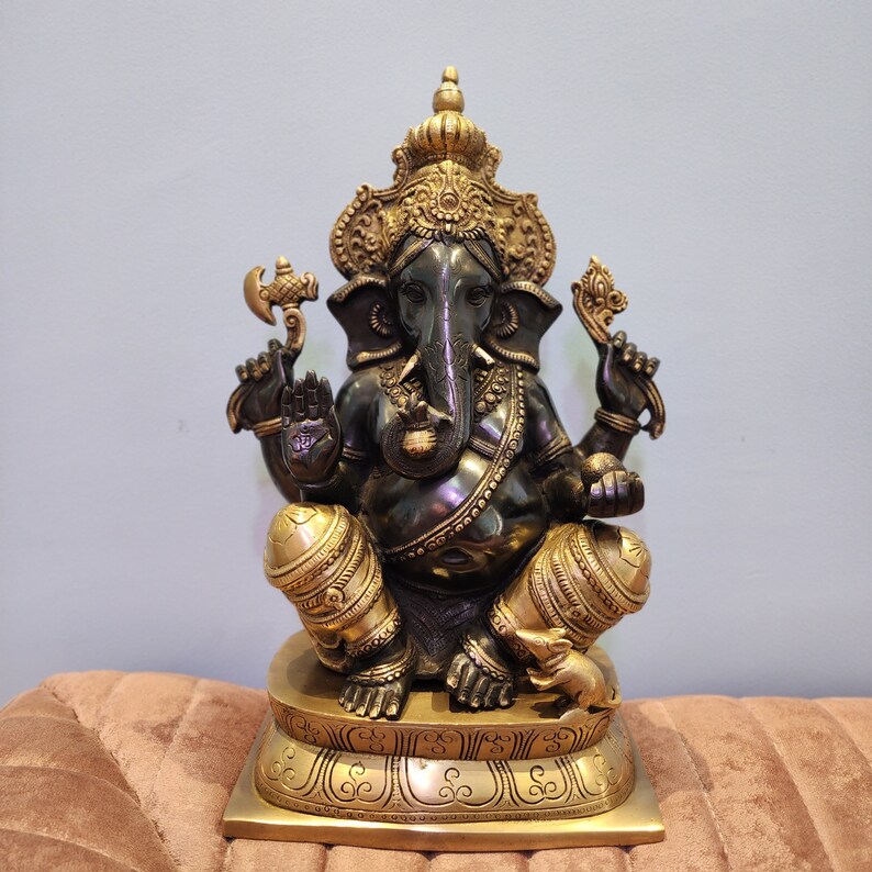 May include: A gold and black metal statue of Ganesha, the Hindu god of wisdom and prosperity, sitting on a decorative base. The statue is intricately detailed and features a traditional headdress and a trunk.