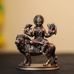 Könnte beinhalten: Eine Bronzestatue einer hinduistischen Göttin, die auf einem Löwen reitet. Die Göttin ist mit mehreren Armen dargestellt, die verschiedene Waffen und Symbole halten. Sie trägt ein farbenfrohes Sari und Schmuck. Der Löwe steht auf einem runden Sockel.